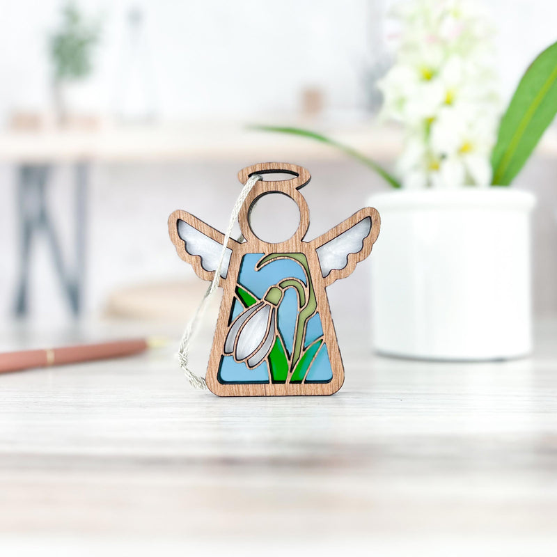 The Forged Flare® Snowdrop Ornament, a 3.5" Mother's Angels® figurine with stained glass wings and a snowdrop birth flower design, stands on light wood with a blurred white pot and plant in the background.