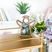 The Forged Flare® Snowdrop Ornament (Mother’s Angels®, 3.5” H angel figurine) with stained glass details is displayed on a gold shelf, backed by jars of flowering plants, including snowdrops, and books on white shelves.