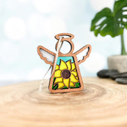 A Forged Flare® Mother's Angels® Sunflower Ornament 3.5" angel figurine with stained glass-style wings stands on a wooden surface, framed by a plant and soft blue background—listed as an Amazon Warehouse Return.