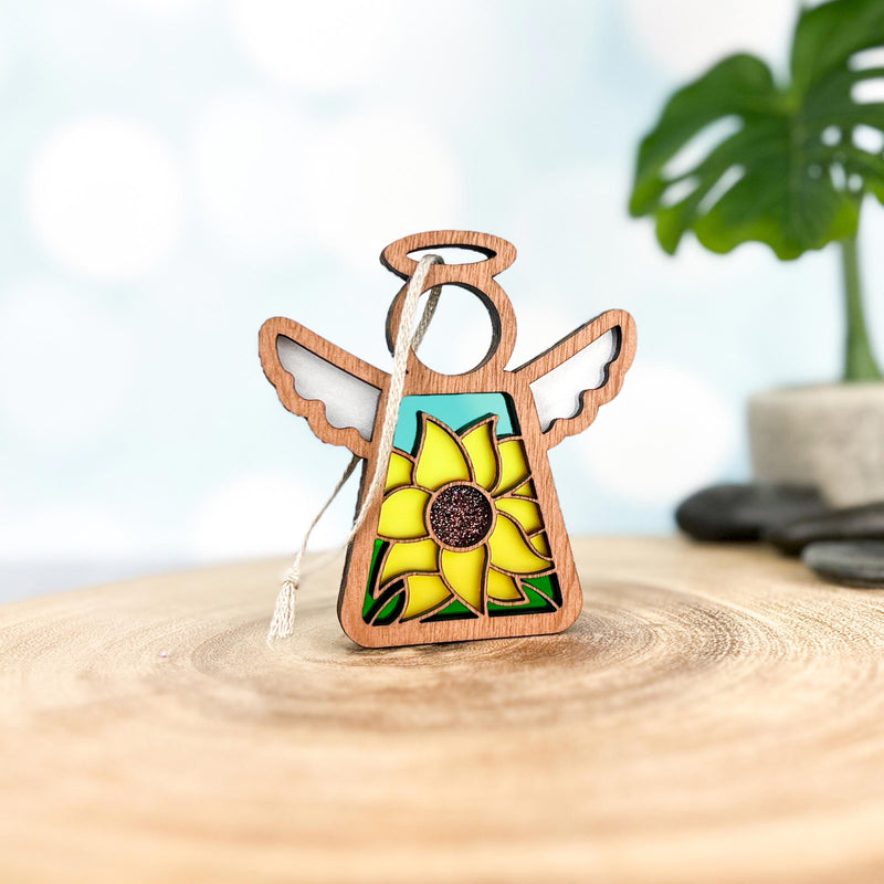 A Forged Flare® Mother's Angels® Sunflower Ornament 3.5" angel figurine with stained glass-style wings stands on a wooden surface, framed by a plant and soft blue background—listed as an Amazon Warehouse Return.
