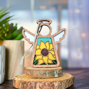 A Forged Flare® Mother's Angels® Sunflower Ornament, a 3.5" wooden angel figurine with stained-glass wings, stands on tree slices; blurred plants and warm lights in the background evoke coziness from Amazon warehouse return inventory.