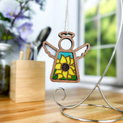 A Forged Flare® Mother's Angels® Sunflower Ornament, a 3.5" stained glass-style angel figurine marked as a warehouse return, hangs from a silver stand on a wooden table with blurred flowers and an inventory organizer in the background.