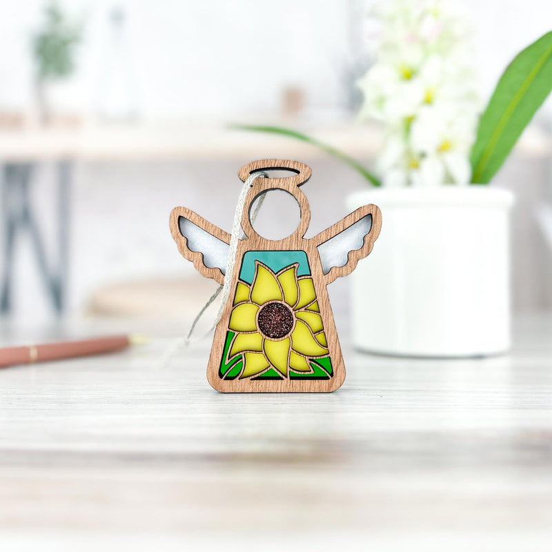 This Forged Flare® Mother's Angels® Sunflower Ornament is a 3.5" wooden angel figurine shown on a light wood surface beside a potted plant, with the background softly blurred.