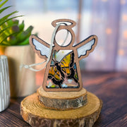 The Forged Flare® Mother's Angels® Swallowtail Butterfly Ornament features a 3.5" wooden angel with white wings and a stained glass butterfly, displayed on a tree slice stand with a white string. Potted plant decor accents the wooden surface nearby.