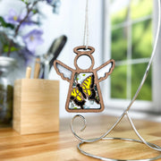 A Forged Flare® Mother's Angels® Warehouse Return - Swallowtail Butterfly Ornament (3.5" H angel figurine) hangs from a silver stand on a wooden table, surrounded by flowers and soft natural light from a window.