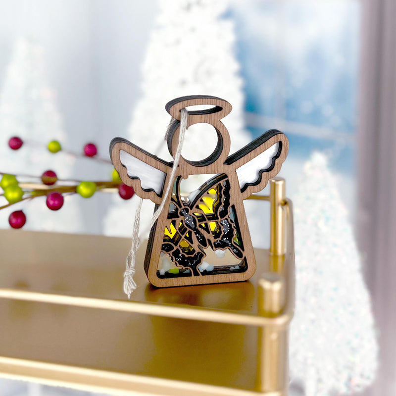 A Forged Flare® Mother's Angels® Swallowtail Butterfly Angel Ornament (3.5") with white wings and butterfly cutout rests on a gold shelf, backed by blurred white Christmas trees and festive red and green berries.