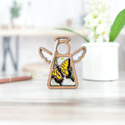 A Forged Flare® Mother's Angels® 3.5" Swallowtail Butterfly Ornament stands on a light table next to a white potted plant, with softly blurred shelves and décor in the background.