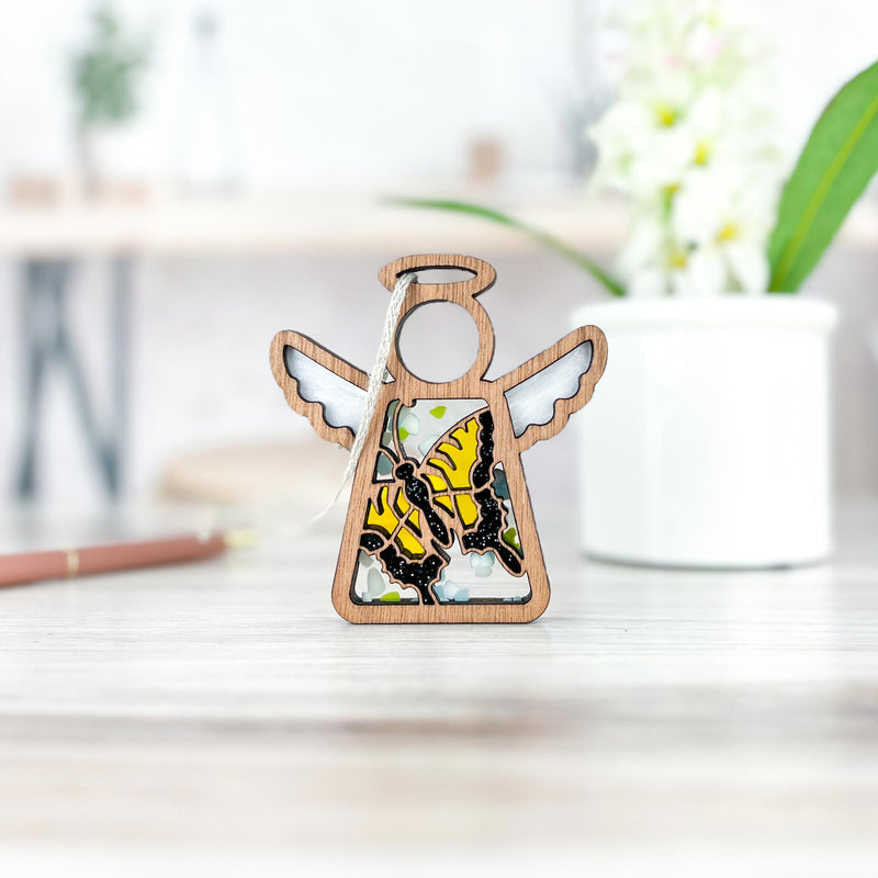 A Forged Flare® Mother's Angels® 3.5" Swallowtail Butterfly Ornament stands on a light table next to a white potted plant, with softly blurred shelves and décor in the background.