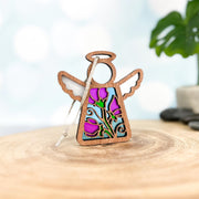 The Sweet Pea Ornament, a 3.5" wooden angel figurine by Forged Flare® Mother's Angels®, features white wings and a sweet pea flower design, with silver string attached, standing on a wooden surface against a blurred background.