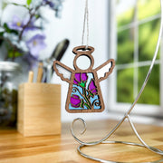 The Forged Flare® Mother's Angels® Sweet Pea Ornament, a 3.5" wooden angel figurine with a stained glass-style sweet pea flower, hangs from a silver stand on a wooden table, surrounded by blurred flowers and art supplies near a window.