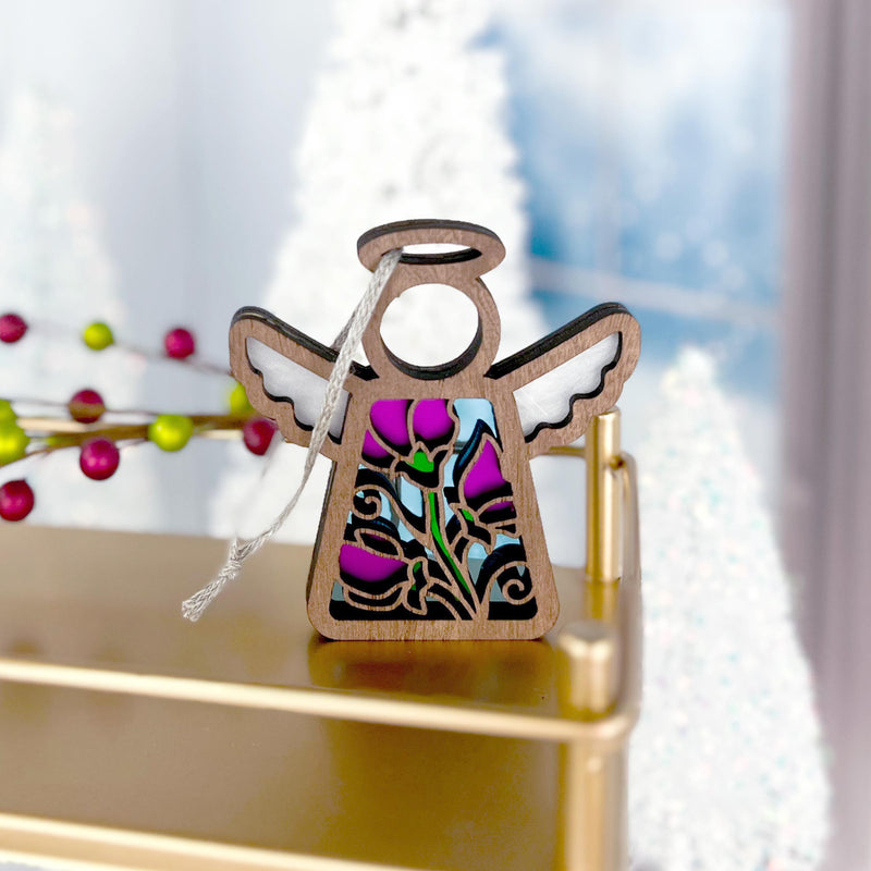 The Forged Flare® Mother's Angels® Sweet Pea Ornament, a 3.5" stained glass-style angel with pink and purple sweet pea flowers for June, sits on a gold shelf with blurred white Christmas trees and berry décor in the background.
