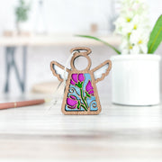 The Sweet Pea Ornament, a 3.5" angel figurine by Forged Flare® Mother's Angels®, features stained-glass-style sweet pea flowers, April’s birth flower, and stands on a light wood surface with blurred flowers and a pencil in the background.