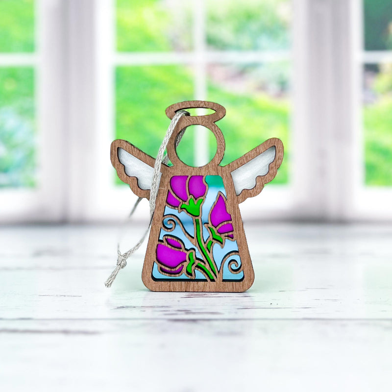 The Forged Flare® Mother's Angels® Sweet Pea Ornament is a 3.5" wooden angel figurine featuring a stained glass-style panel with sweet pea flowers and green leaves, shown on a white surface with large windows and greenery in the blurred background.