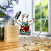The Teacher Ornament with Imperfections by Forged Flare®, a 3.5" Mother's Angels® wooden angel with a stained glass apple and pencils, hangs from a metal stand on a desk, flowers and window blurred in the background.