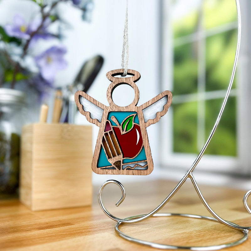 The Teacher Ornament with Imperfections by Forged Flare®, a 3.5" Mother's Angels® wooden angel with a stained glass apple and pencils, hangs from a metal stand on a desk, flowers and window blurred in the background.