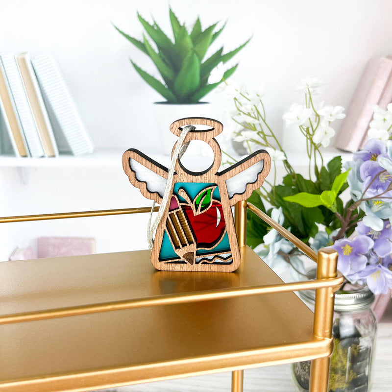 A Forged Flare® Mother's Angels® Teacher Ornament with Imperfections, a 3.5" handcrafted angel figurine, hangs on a gold shelf with purple and white flowers in vases and stacked books on white shelves behind.