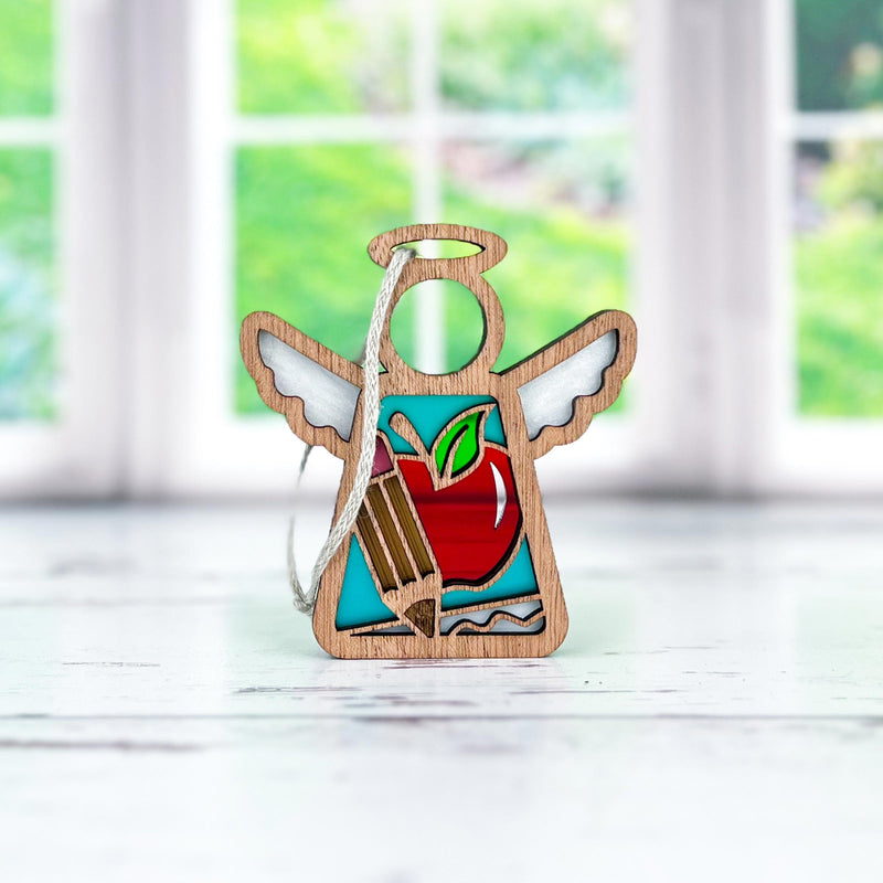 The Teacher Ornament with Imperfections is a 3.5" angel figurine from Forged Flare® Mother's Angels®, displayed on a light surface with red apple and pencil details. Blurred green scenery appears through the window in the background.