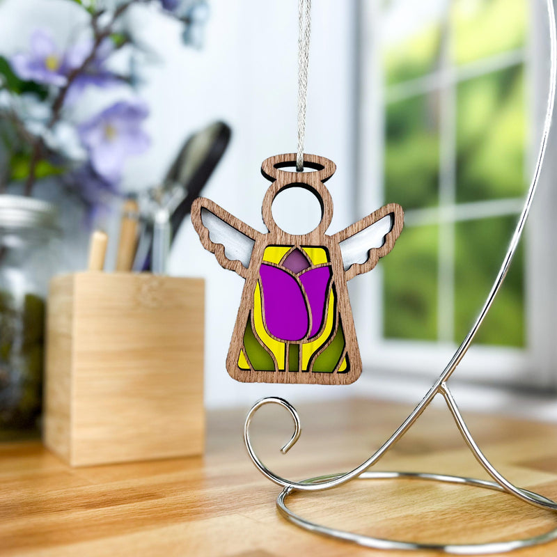 The Tulip Ornament, a 3.5" angel figurine from Mother's Angels® by Forged Flare®, with purple and yellow accents, hangs beautifully from a stand. The background features a softly blurred indoor scene with natural light streaming through a window and several decorative items adorning a wooden surface, making it the perfect addition to any Christmas tree.