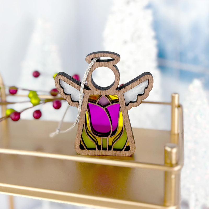 The Forged Flare® Tulip Ornament | 3.5" Angel Figurine from Mother's Angels® features stained glass-style purple, green, and pink accents, hanging by a string on a gold shelf amid festive berries and snowy decor with tulip motifs blurred behind.