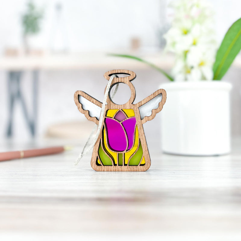 The Forged Flare® Mother's Angels® Tulip Ornament is a 3.5" wooden angel figurine with wings and a halo, featuring a pink and yellow stained glass-style tulip. It’s displayed on a white surface with a blurred plant in the background.