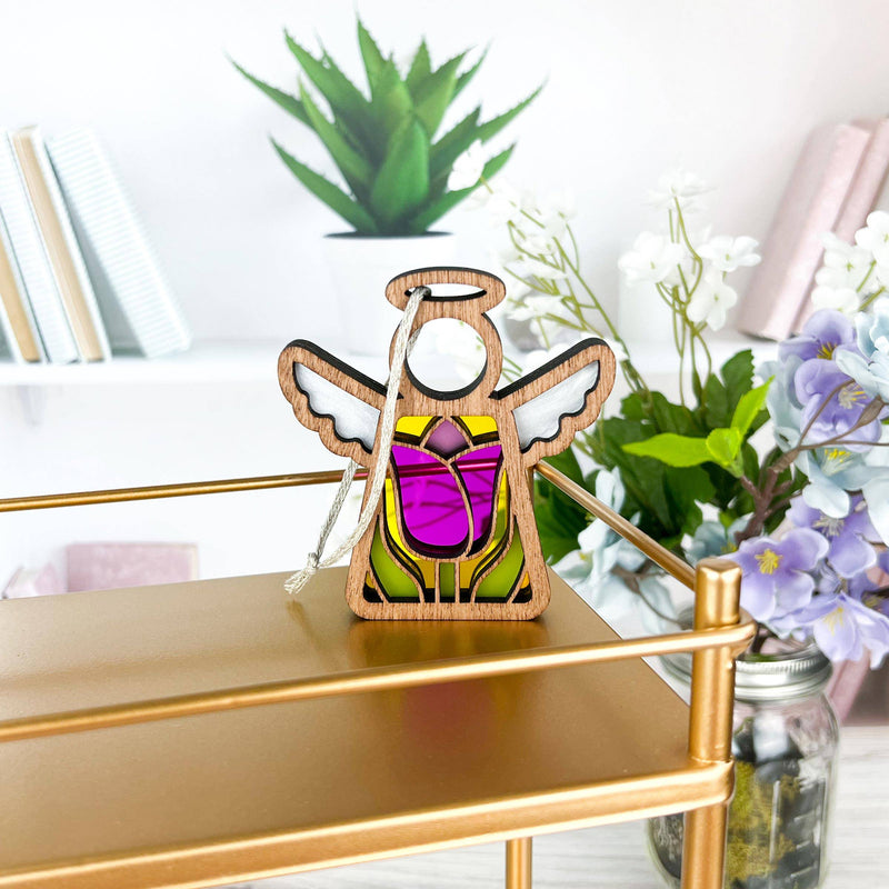 A small wooden angel figurine with stained glass-like wings and robe, named the 3.5" Angel Figurine from Mother's Angels® by Forged Flare®, sits on a gold shelf. Behind it are potted plants and books on a white shelf, along with the Tulip Ornament. The background includes white walls and a jar of purple flowers.