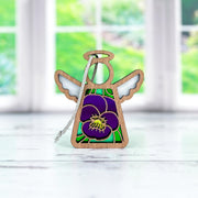The Violet Ornament by Forged Flare® | 3.5" Mother's Angels® Angel Figurine is a charming small wooden ornament featuring purple and green stained glass-style wings and body, adorned with a violet flower with a yellow center on its front. Placed on a white surface, large windows showcasing a green outdoor scene can be seen in the background. Perfect as a birthday gift, it adds charm to any setting.