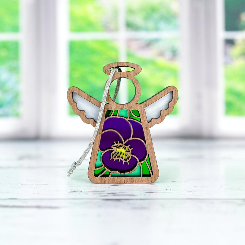 The Violet Ornament by Forged Flare® | 3.5" Mother's Angels® Angel Figurine is a charming small wooden ornament featuring purple and green stained glass-style wings and body, adorned with a violet flower with a yellow center on its front. Placed on a white surface, large windows showcasing a green outdoor scene can be seen in the background. Perfect as a birthday gift, it adds charm to any setting.