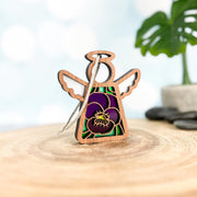 The Forged Flare® Violet Ornament with Imperfections is a 3.5" Mother's Angels® figurine featuring stained glass-style wings and a violet pansy design, hanging by a string on wood with a blurred plant in the background.