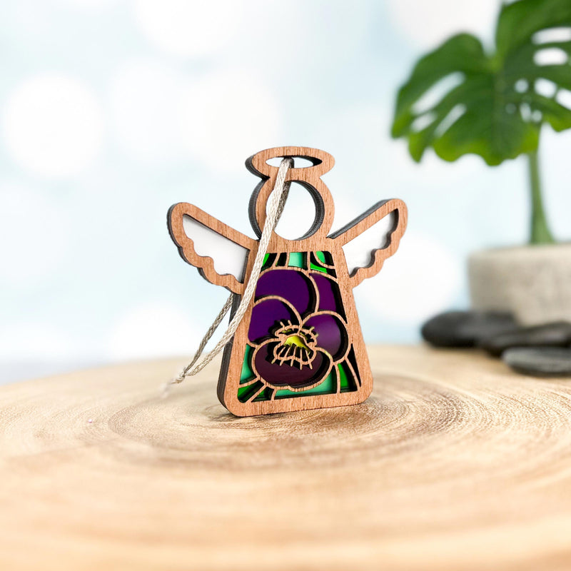 The Forged Flare® Violet Ornament with Imperfections is a 3.5" Mother's Angels® figurine featuring stained glass-style wings and a violet pansy design, hanging by a string on wood with a blurred plant in the background.