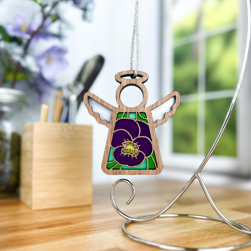 The Forged Flare® Mother's Angels® Violet Ornament with Imperfections is a 3.5" angel figurine, featuring a purple flower design, displayed on a silver stand against a bright room with window, pens, and a blurred violet centerpiece.