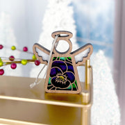 The Forged Flare® Violet Ornament | 3.5" (H) Mother's Angels® Angel Figurine, featuring stained glass-style wings and a purple-green dress, rests on gold with festive white tree decorations and red berries in the background.