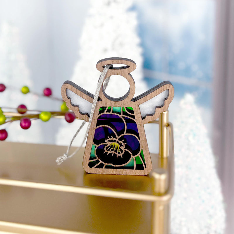 The Forged Flare® Violet Ornament | 3.5" (H) Mother's Angels® Angel Figurine, featuring stained glass-style wings and a purple-green dress, rests on gold with festive white tree decorations and red berries in the background.
