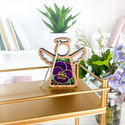 A 3.5" Forged Flare® Mother's Angels® Violet Ornament with Imperfections hangs on a gold shelf, surrounded by books, a potted plant, and a vase with white and purple flowers in the background.