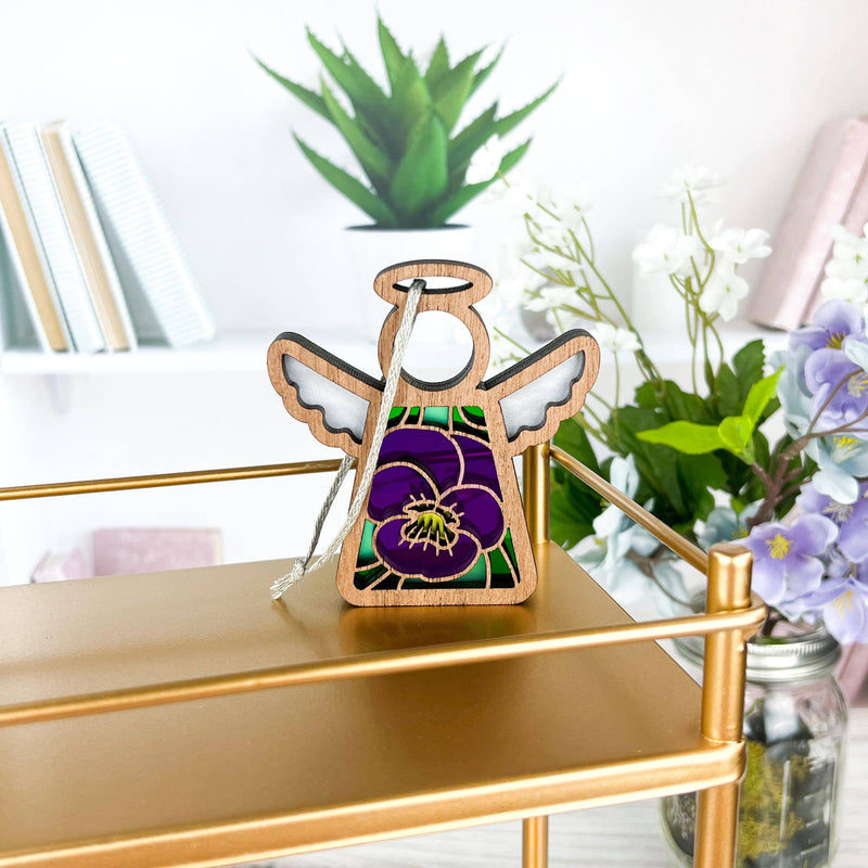 A 3.5" Forged Flare® Mother's Angels® Violet Ornament with Imperfections hangs on a gold shelf, surrounded by books, a potted plant, and a vase with white and purple flowers in the background.