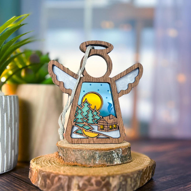 The Forged Flare® Warm Welcome Ornament with Imperfections is a 3.5" Mother's Angels® handcrafted wooden angel figurine featuring an open center with trees, a moon, and a log cabin scene, standing on a wood slice.