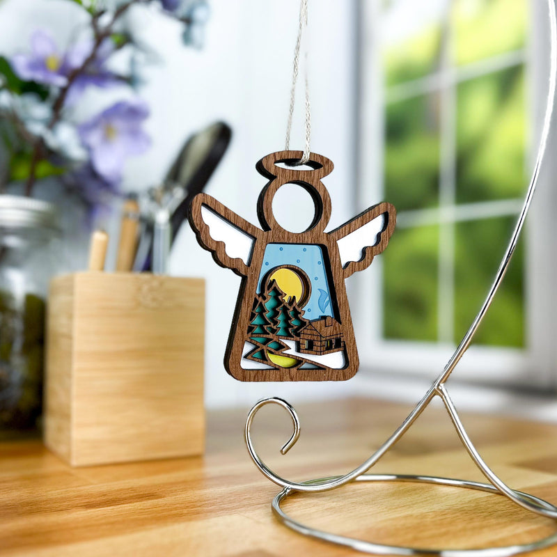 The Warm Welcome Ornament from Forged Flare® features a 3.5" wooden angel figurine with an intricate cut-out design that reveals a scenic landscape of trees, a log cabin, and a full moon. Ideal for enhancing your Christmas tree ornament collection, this Mother's Angels® piece hangs elegantly from a string on a metal stand. The backdrop of blurred greenery and purple flowers hints at an indoor setting near a window.