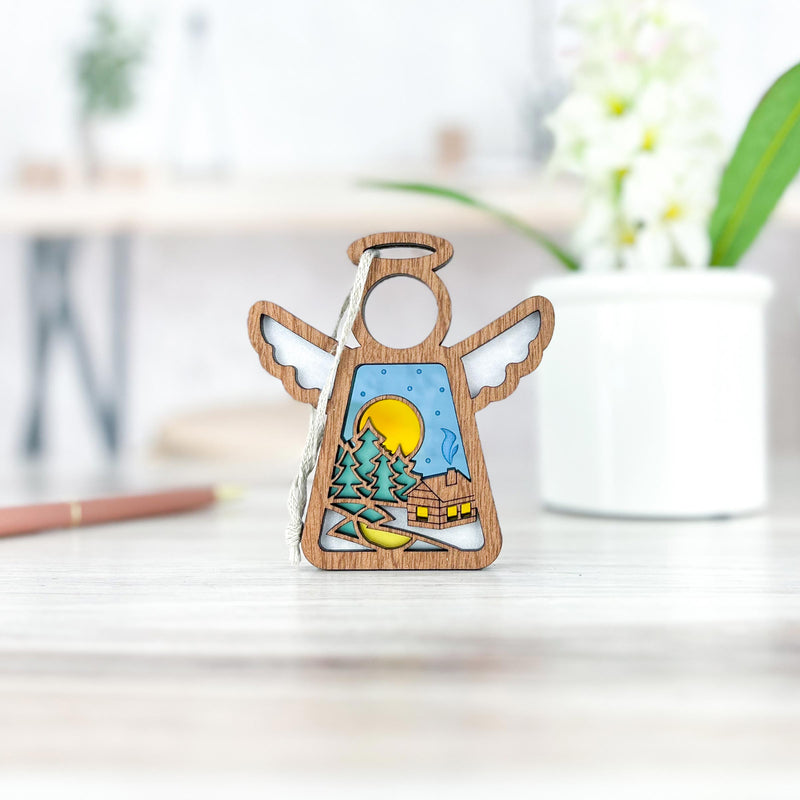 The Warm Welcome Ornament with Imperfections by Forged Flare® (Mother's Angels®) is a 3.5" handcrafted angel figurine featuring a painted log cabin, trees, and yellow moon—displayed on a light wood table with blurred flowers and a pencil in the background.