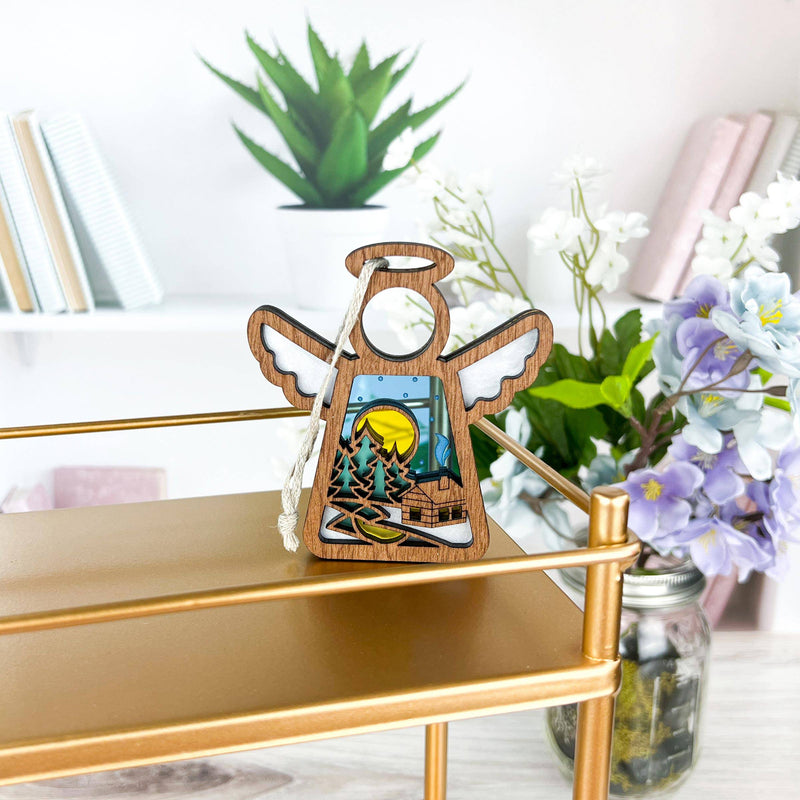 The Warm Welcome Ornament by Forged Flare® is a 3.5" Mother's Angels® figurine with a nature scene, displayed on a gold shelf among books, a potted plant, and flowers—a charming setting for showcasing Christmas ornaments.