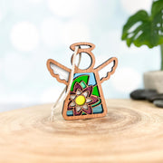 The Forged Flare® Mother's Angels® Water Lily Ornament is a 3.5" wooden angel figurine with stained glass-style wings and a vibrant birth flower design, displayed on wood with a blurred plant and stones in the background.