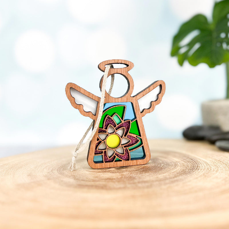 The Forged Flare® Mother's Angels® Water Lily Ornament is a 3.5" wooden angel figurine with stained glass-style wings and a vibrant birth flower design, displayed on wood with a blurred plant and stones in the background.