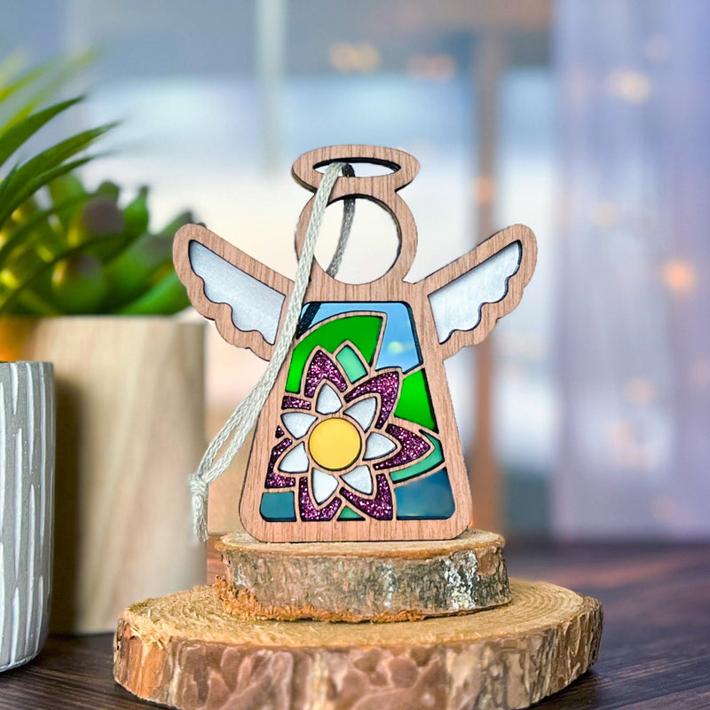 The Forged Flare® Water Lily Ornament | 3.5" (H) Angel Figurine from Mother's Angels® features stained glass-style wings and a vibrant water lily birth flower, standing on wooden slices with softly lit indoor plants in the background.
