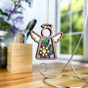 The Forged Flare® Mother's Angels® Water Lily Ornament, a 3.5" wooden angel figurine with stained glass-style wings, hangs from a silver stand on a wooden desk by a blurred window and flowers.