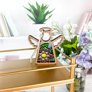 The Water Lily Ornament, a 3.5" angel figurine from Forged Flare® Mother's Angels®, featuring floral stained glass details, hangs on a gold shelf with books, green plants, and purple and white flowers in the background.