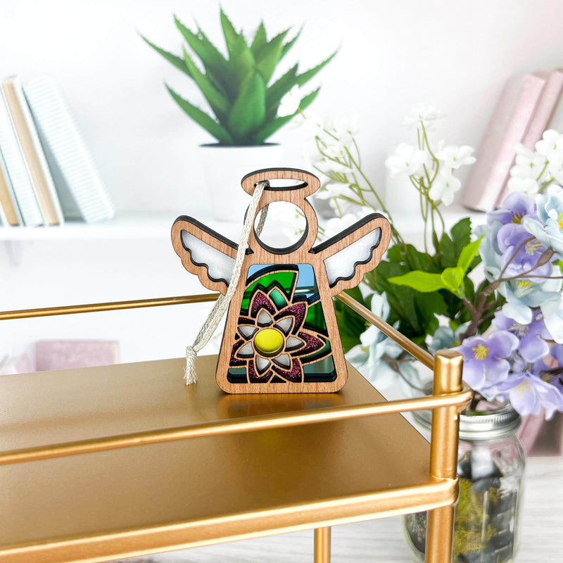 The Water Lily Ornament, a 3.5" angel figurine from Forged Flare® Mother's Angels®, featuring floral stained glass details, hangs on a gold shelf with books, green plants, and purple and white flowers in the background.