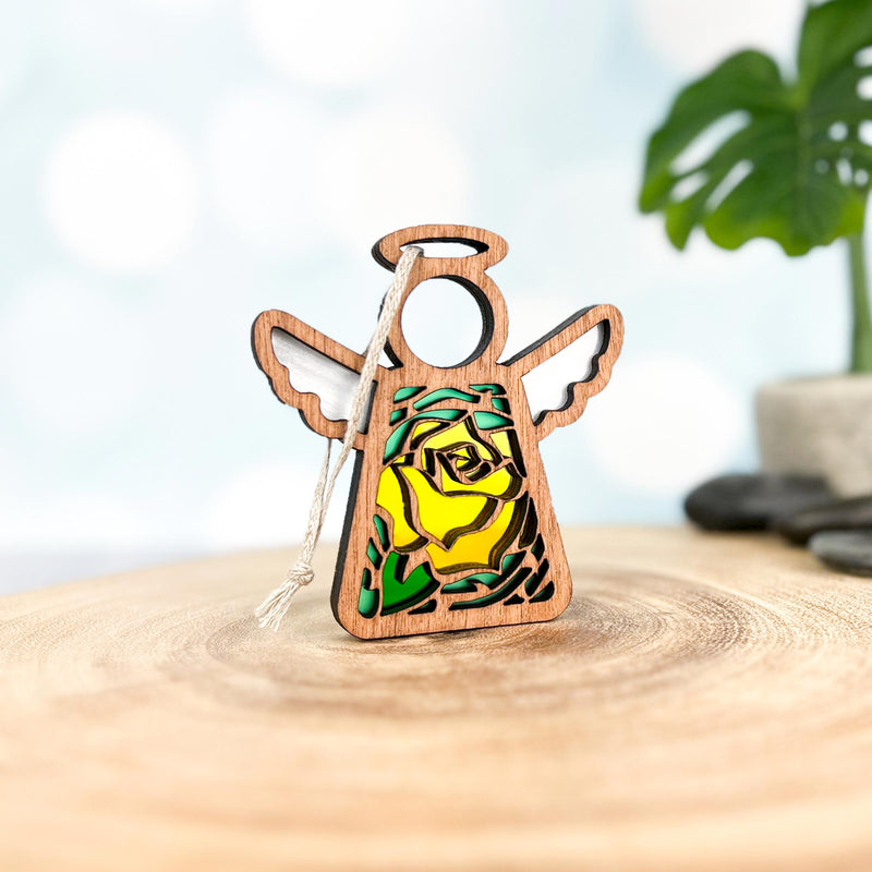 The Forged Flare® Yellow Rose Ornament is a 3.5" Mother's Angels® figurine with outstretched wings, halo, and a stained-glass style yellow rose—the June birth flower—at its center. Shown on wood with blurred plants and stones behind.