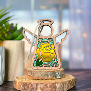 The Forged Flare® Mother's Angels® Yellow Rose Ornament is a 3.5" wooden angel figurine with a stained glass yellow rose design, symbolizing June’s birth flower, displayed on a tree slice with a plant and softly blurred background.