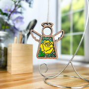 The Forged Flare® Yellow Rose Ornament—a 3.5" Mother's Angels® figurine with white wings and a yellow rose—hangs from a silver stand on a wooden desk, blurred plants and a window in the background.