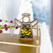 The Forged Flare® Yellow Rose Ornament, a 3.5" Mother's Angels® angel figurine featuring June’s birth flower, sits on a gold surface with snowy trees and red berries in the festive background.
