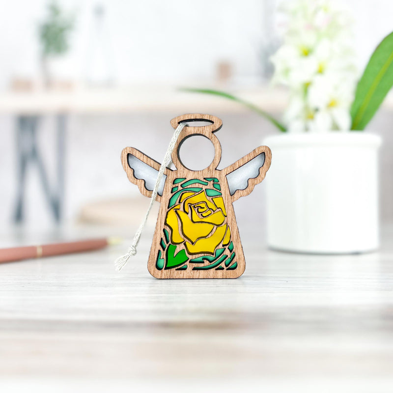 A Yellow Rose Ornament, a 3.5" angel figurine by Forged Flare's® Mother's Angels® collection, features intricately carved wooden wings and a torso adorned with a yellow rose birth flower design. It stands on a light-colored wooden surface with a white string attached to its head. The background includes a blurred white pot with flowers and a pencil.