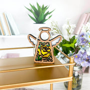 The Forged Flare® Yellow Rose Ornament, a 3.5" Mother's Angels® figurine with June’s birth flower, stands on a gold shelf with books, green plants, and flowers in the background.